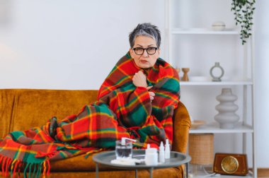 Yaşlı kadın üşütmüş ya da grip olmuş, battaniyeye sarılmış ve yakınlarda ilaç alıyormuş.