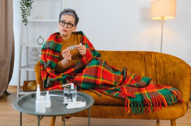 Yaşlı kadın kendini iyi hissetmiyor, kanepeye sıcak bir battaniyeye sarılmış, hastalık ve rahatlama için haplar tutuyor.