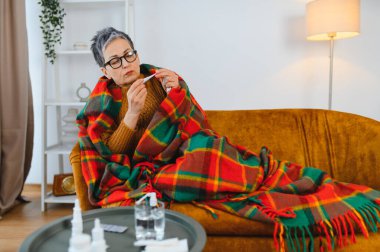 Kanepede oturan olgun bir kadın kendini iyi hissetmiyor ve ateşini kontrol ediyor.