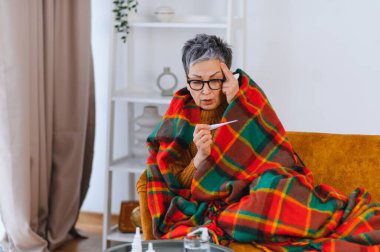 Yaşlı kadın soğuk algınlığı ve gripten rahatsız, elinde tıbbi termometreyle alnına dokunuyor, evdeki bir kanepede dinleniyor.