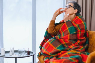 Soğuk algınlığı ya da nezle olan yaşlı bir kadın, evinde sıcak bir battaniyeye sarılmış, ferahlamak için burun spreyi kullanıyor.