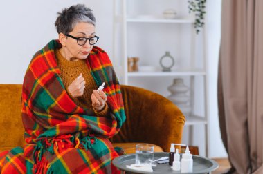 Soğuk kanlılıkla kanepede oturan, sıcak battaniyeye sarılı yaşlı kadın, rahatlamak için burun damlası kullanmaya hazırlanıyor.