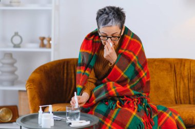 Yaşlı kadın grip belirtilerinden rahatsız, sıcak bir battaniyeye sarılıp burnunu siliyor, ilaç alıyor.