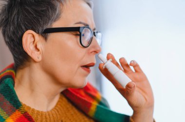 Olgun bir kadın gözlük ve eşarp takıyor, burnuna burun spreyi sürüyor, soğuk algınlığı veya alerji semptomları ve tıkanıklığı kontrol ediyor.