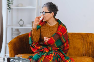 Soğuk algınlığı ve grip semptomlarını yatıştırmak için burun spreyi kullanan kanepedeki son sınıf öğrencisi kadın, evde dinlenme sırasında battaniyenin altında rahat.