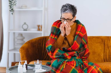 Yaşlı kadın ekose battaniyeye sarılmış, burnunu mendille siliyor. İlaçlar ve su masada.