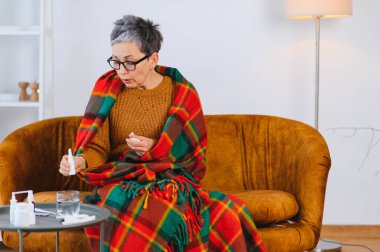 Yaşlı kadın soğuk algınlığı ya da nezle yüzünden kendini kötü hissediyor, battaniyeye sarılmış, kanepede burun spreyi ve mendil kullanıyor.