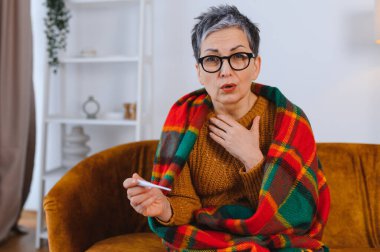 Hasta kıdemli bir kadın evdeki kanepede kendini kötü hissediyor. Soğuk algınlığı, grip ve boğaz ağrısı var. Termometresi sıkıntılı görünüyor.