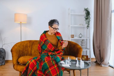 Sıcak bir battaniyeye sarılı, kanepede oturmuş termometreye bakan, hastalıklı ve ateşli yaşlı bir kadın.