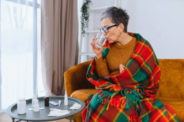 Yaşlı kadın sıcak bir battaniyeyle üstünü örtüyor, su içiyor ve evde hastalığını tedavi etmek için ilaç alıyor.