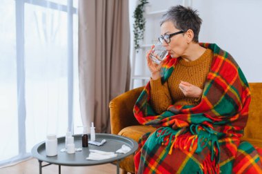 Olgun bir kadın su içiyor ve battaniyeye sarılı bir şekilde hap alıyor. Masadaki ilaçlarla bir kanepede dinleniyor.