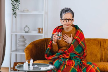 Bir kadın sıcak bir battaniyeye sarılmış, kanepeye oturmuş ve elinde bir bardak su, masada ilaç tutarken kendini kötü hissediyor.