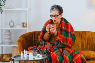 Son sınıf öğrencisi kadın battaniyeye sarılmış, sıcak çayını yudumlarken bir kanepede dinleniyor, rahatlamak için ilaçlarla çevrili.