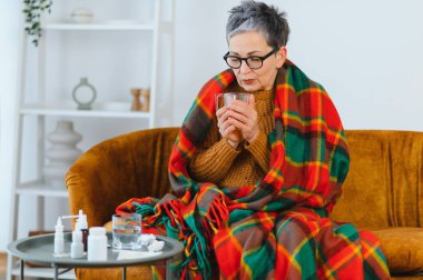 Yaşlı kadın, battaniyeye sarılı, soğuk algınlığı ya da gripten hasta hissediyor. Masasında sıcak içecek ve ilaç tutuyor.
