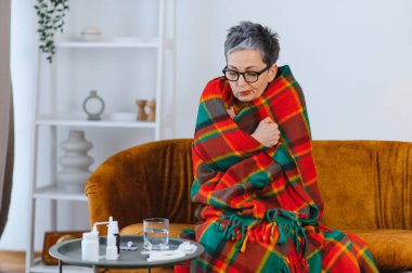 Yaşlı kadın grip semptomlarıyla kendini kötü hissediyor, kanepeye oturmuş, ekose bir battaniyeye sarılmış, evinde üşüyor ve titriyor.