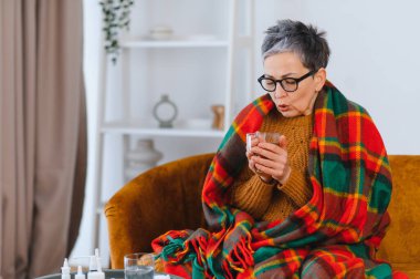 Kanepeye oturmuş, sıcak battaniyeye sarılmış, rahatlamak için sıcak çay fincanına üfleyerek hasta olan olgun bir kadın.