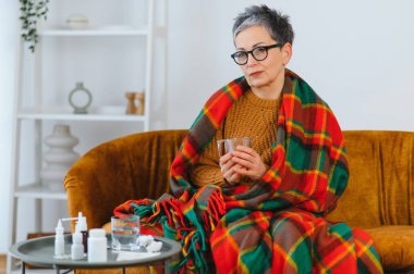 Olgun bir kadın kendini hasta ve iyi hissetmiyor, battaniyeye sarılmış bir şekilde kanepeye oturuyor ve grip belirtileri için ilaç alıyor.