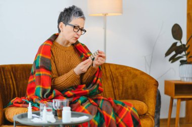 Yaşlı bir kadın hastalıktan muzdarip, kanepeye battaniyeye sarılmış, ilaç paketinde tıbbi bilgiler okuyor.