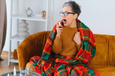 Kendini hasta hisseden bir kadın, battaniyeye sarılmış, boğaz spreyi kullanıyor ya da soğuk algınlığı belirtileri gösteriyor.