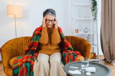 Yaşlı bir kadın kronik baş ağrısı ya da migren geçiriyor. Battaniyeye sarılmış bir şekilde oturuyor. Yanında da ilaç ve su var.
