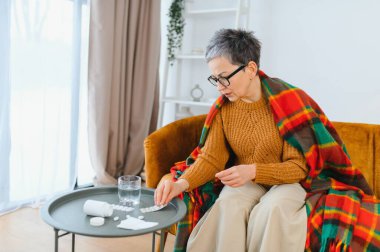Kanepeye kareli battaniyeye sarılmış, evde dinlenirken baş ağrısı veya grip ilacı alan olgun bir kadın, düşünceli görünüyor.