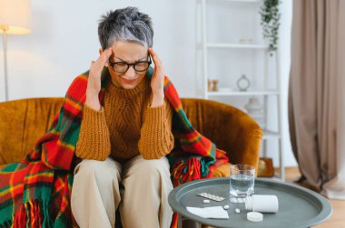 Yaşlı kadın şiddetli baş ağrısı ya da migren geçiriyor, battaniyeye sarılmış ve masada su ile oturuyor.
