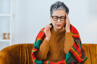 Baş ağrısı ya da migrenden muzdarip yaşlı bir kadın, evde battaniyeye sarılı dinlenirken kendini rahatsız hissediyor ve tapınakları tutuyor.