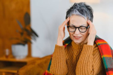 Baş ağrısı ve migrenden muzdarip yaşlı bir kadın, gözlük ve sıcak bir battaniye takıyor, evde rahatsızlık ve stres yaşıyor.