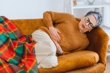 Üst düzey bir kadın karın ağrısı çekiyor. Battaniyesiyle kanepede yatarken karnını tutuyor.