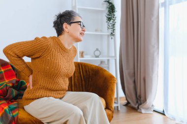 Son sınıf öğrencisi kadın yoğun bel ağrısı çekiyor. Ev ortamında bir kanepede otururken belini tutuyor.