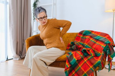 Yaşlı bir kadın koltukta otururken bel ağrısı çekiyor. Rahatsızlık ve sağlık sorunları yaşıyor.