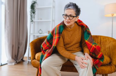 Gri saçlı, gözlüklü ve ekose battaniyeli yaşlı bir kadın, evinde diz ağrısından rahatsız oluyor.