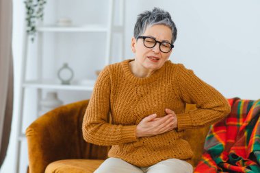 Olgun bir kadın ağır kalp ağrısı ya da göğüs rahatsızlığı yaşıyor, karnını tutuyor, evde oturuyor. Kalp sağlığı sorunları kavramı