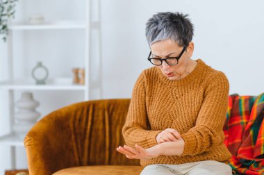Evde yaşlı bir kadın, tahriş olmuş cildini inceliyor, kolunu kaşıyor ve alerjik reaksiyondan rahatsız oluyor.