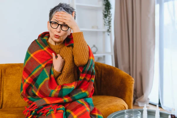 Ateşli ve baş ağrısı olan yaşlı kadın evdeki kanepede battaniyeye sarılmış, dinleniyor ve iyileşme sürecinde huzur arıyor.