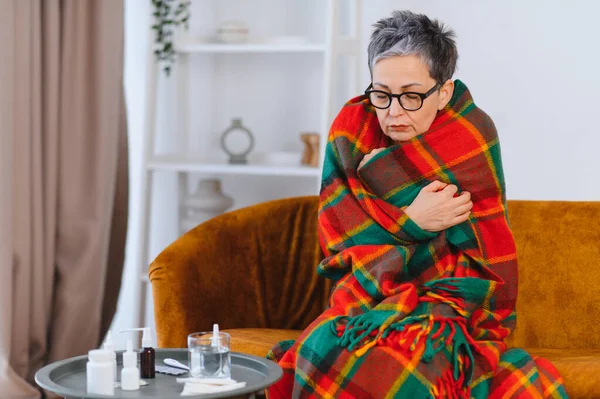 İyi olmayan yaşlı kadın battaniyeye sarılmış, ateşten ya da soğuktan titriyor masada ilaçlarla bir kanepede otururken.