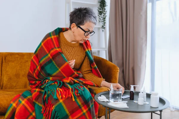 Yaşlı bir kadın soğuk algınlığı ya da gripten muzdarip, kanepeye sarılmış, ilaç ve su için uzanan