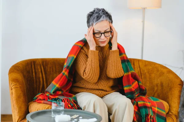 Ağır baş ağrısı çeken yaşlı bir kadın, battaniyeli bir sandalyede otururken elleriyle başını tutuyor.