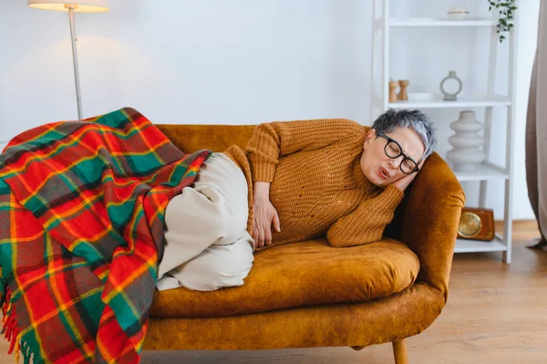 Evde kanepede yatarken mide ağrısı çeken yaşlı bir kadın rahatsızlığı ve rahatsızlığı var.