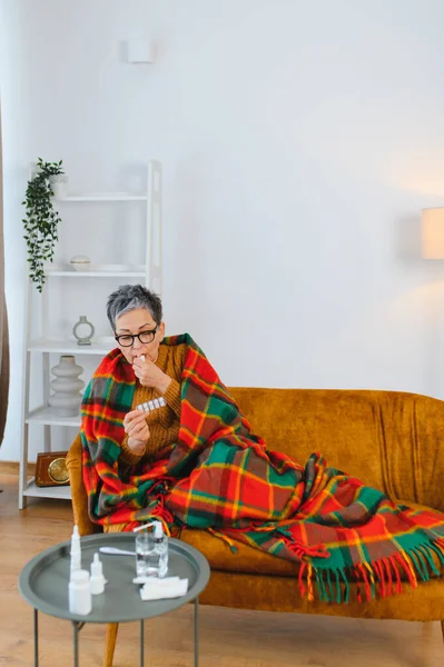 Yaşlı kadın kendini hasta hissediyor, battaniyeye sarılmış, haplar alıyor, sağlığını içeride kontrol ediyor.