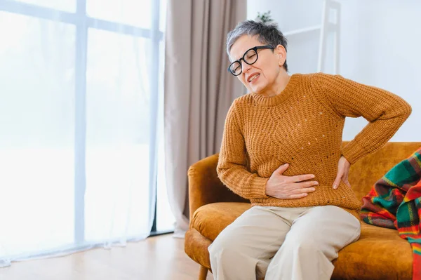 Evde bir kanepede otururken karın ağrısı ve krampları olan yaşlı bir kadın kendini hasta hissediyor.