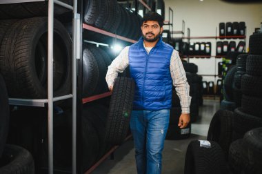 Mavi yelekli Hintli bir adam araba lastiği taşıyor, lastik dükkanından geçiyor, otomotiv ve perakende satış yapıyor.