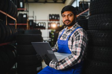 Tamirci olarak çalışan genç Hintli bir adam, tulum giyiyor ve bir lastik servis merkezinde dizüstü bilgisayar kullanıyor.