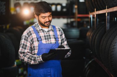 Mavi tulumlu ve ekose gömlekli bir erkek işçi bir lastik dükkanında envanter ve teftiş için bir pano kullanıyor.
