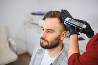 Trikoloğum, klinikte modern teşhis ekipmanlarıyla genç bir erkek hastanın saç ve kafa derisini inceleyip teşhis ediyor.