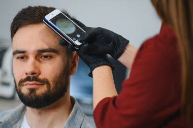 Trikoloğum, teşhis için özel lensli akıllı telefon kullanarak erkek bir hastanın kafa derisini ve saç durumunu inceliyor.