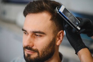 Özel bir cihaz ve akıllı telefon kullanan bir erkek hasta üzerinde saç ve kafa derisi trikolojisi muayenesi yapan bir tıp uzmanı.