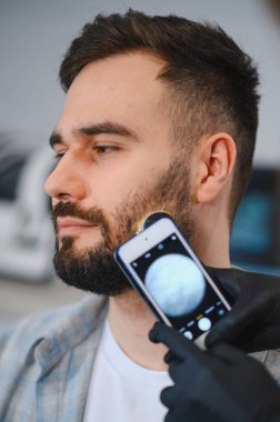 Trikoloğum, bir klinikteki akıllı telefona bağlı modern bir dermatoskopla hastanın sakal kıllarını ve derisini inceliyor.