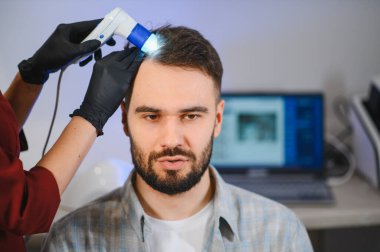 Trikoloğum, klinikte saç kaybı ve kelliği teşhis etmek ve tedavi etmek için trikop kullanarak genç bir adamın kafa derisini inceliyor.