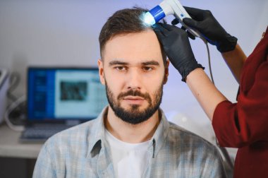 Tıbbi saç muayenesinden geçen bir adam, modern bir klinikte kafa dermatoskobu kullanan bir doktor.
